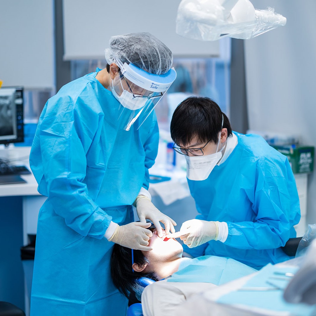 Two dental professionals in blue scrubs and protective gear are performing a dental procedure on a patient lying in a dental chair. The patient is receiving treatment under a bright dental light in a modern clinic setting.